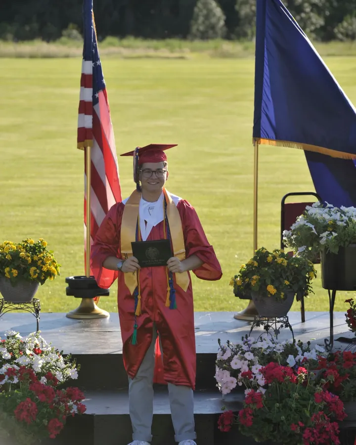 A Project Youth+ graduate in a red gown and gold stole holds a diploma outdoors at commencement